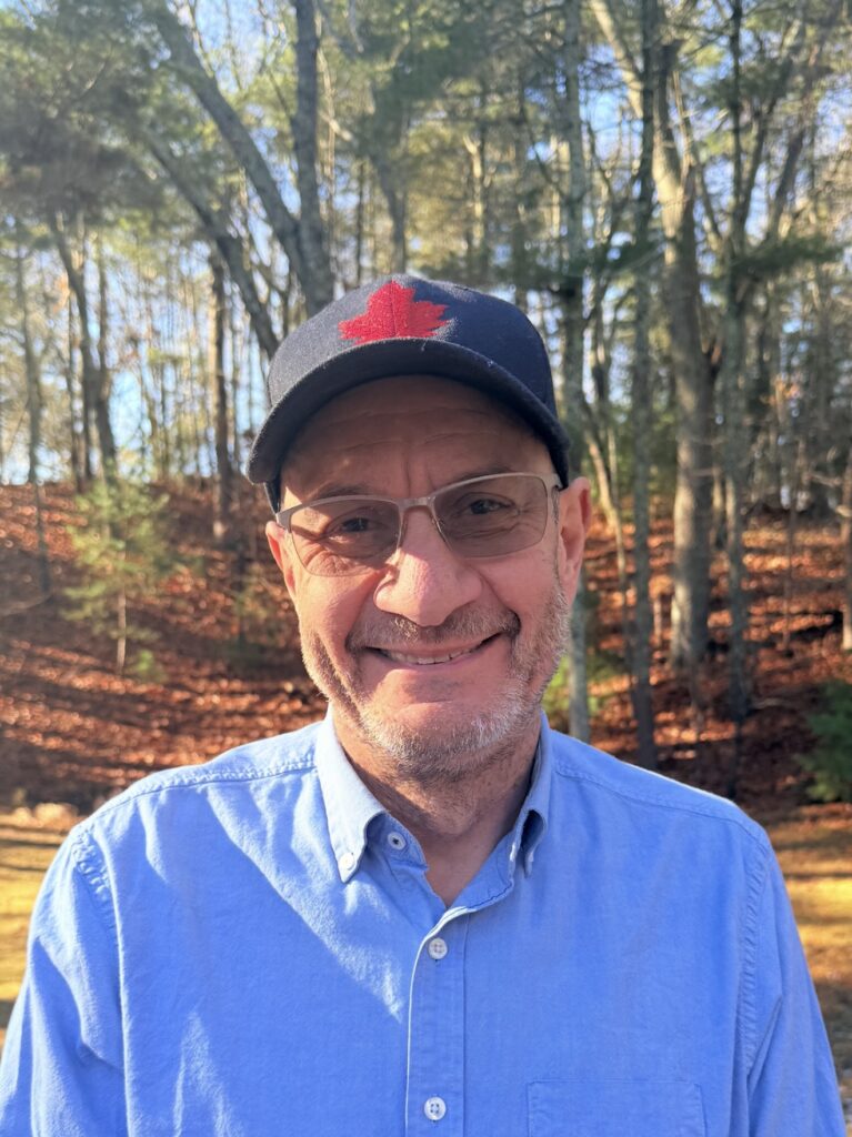 Smiling man in a blue shirt and black cap with a red maple leaf, outdoors in a sunlit forest.